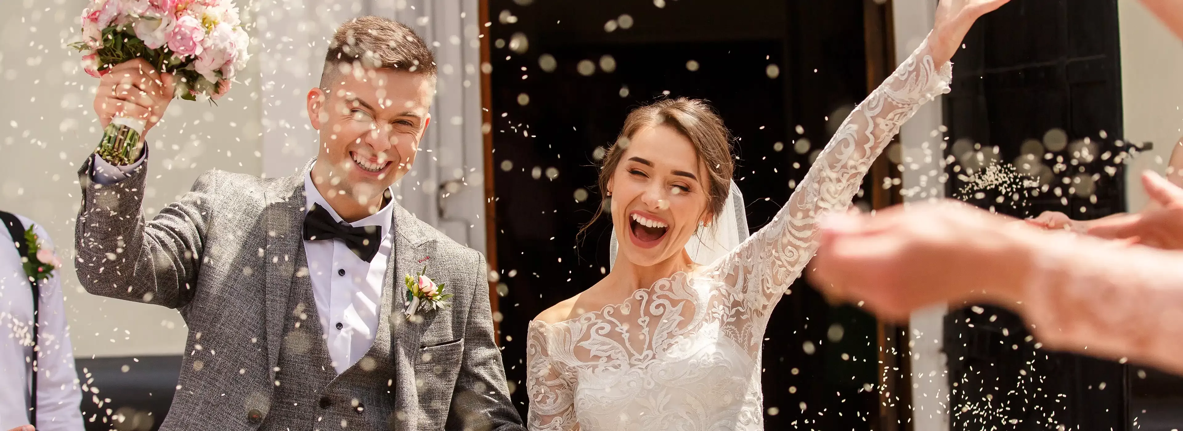 A joyful bride and groom exit a building, smiling and holding hands. The groom is dressed in a gray suit with a bow tie, and the bride wears a white lace gown. Guests shower them with confetti as they celebrate.