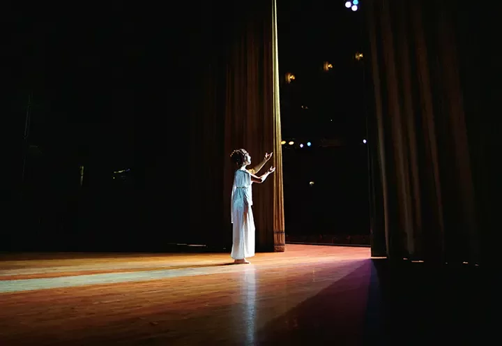 A performer in white stands alone on a dimly lit stage near a curtain, reaching out with both hands toward the light shining from the audience area.