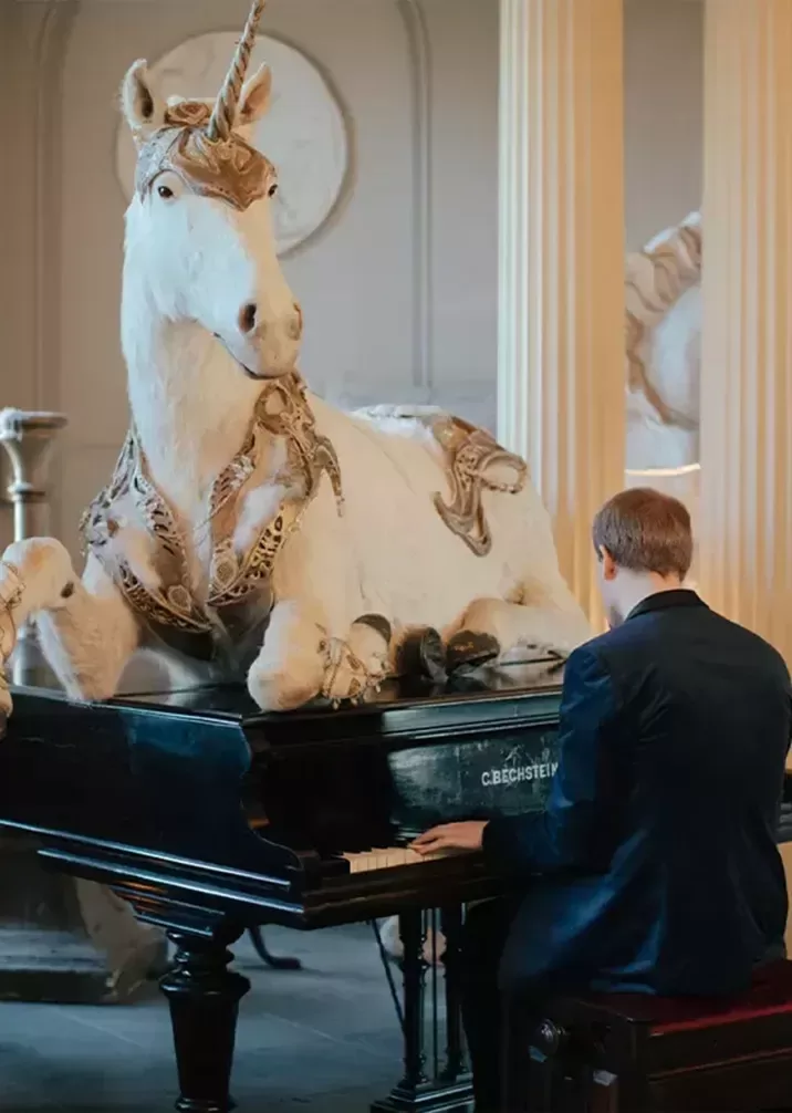 A person in a suit is playing a grand piano, while a large, intricately adorned unicorn statue lies atop the piano. The background features elegant, classical décor with tall columns.