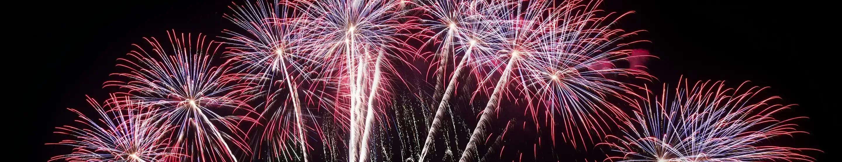 A vibrant fireworks display lights up the night sky over a body of water, with red, white, and blue pyrotechnics creating a stunning visual show. The reflections on the water add to the spectacle.