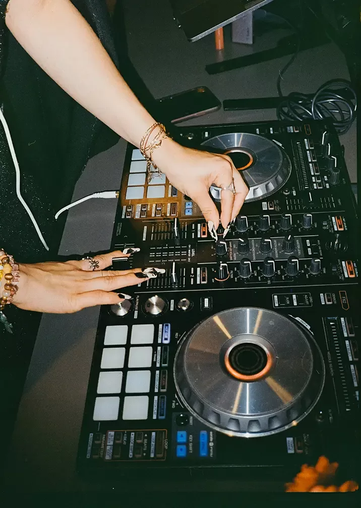 Close-up of a person’s hands adjusting knobs and sliders on a DJ mixer. The person wears several bracelets and rings. The mixer has large silver platters and various controls, with cables connected at the top.
