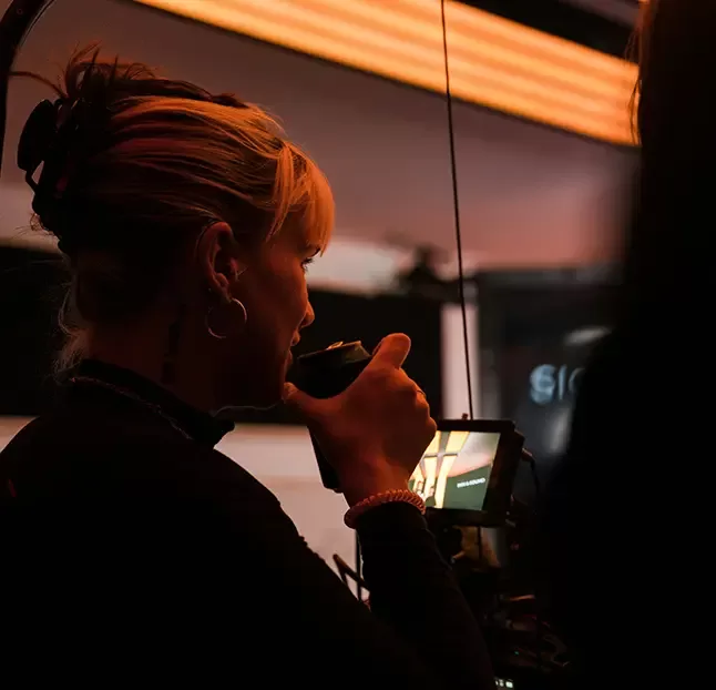 A person with blonde hair and hoop earrings talks into a walkie-talkie, surrounded by dim lighting. They are positioned slightly to the left, holding the device near their mouth. A screen and part of another persons silhouette are visible.