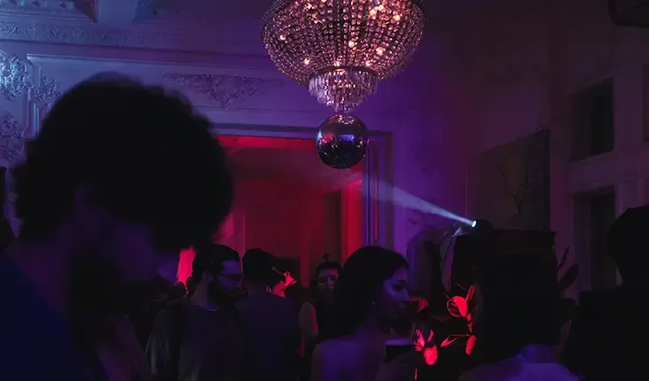 A dimly lit party scene with people silhouetted against dark purple and red lighting. A large chandelier and disco ball are hanging from the ceiling. The atmosphere is vibrant and lively, with attendees mingling and dancing.