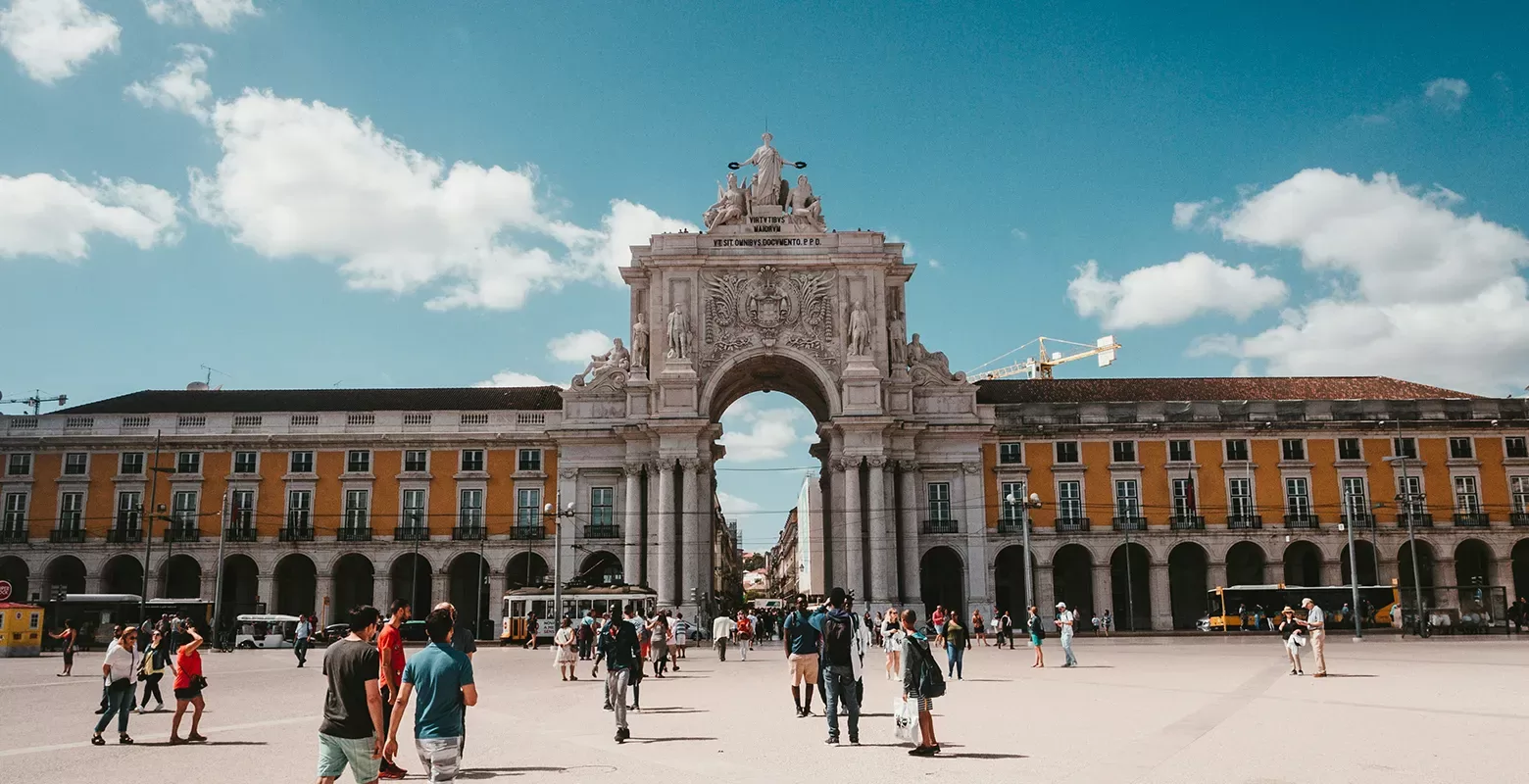 A scenic view of people walking towards a grand building with an arched walkway in Lisbon, showcasing its vibrant culture, historic architecture, and beautiful landscapes.