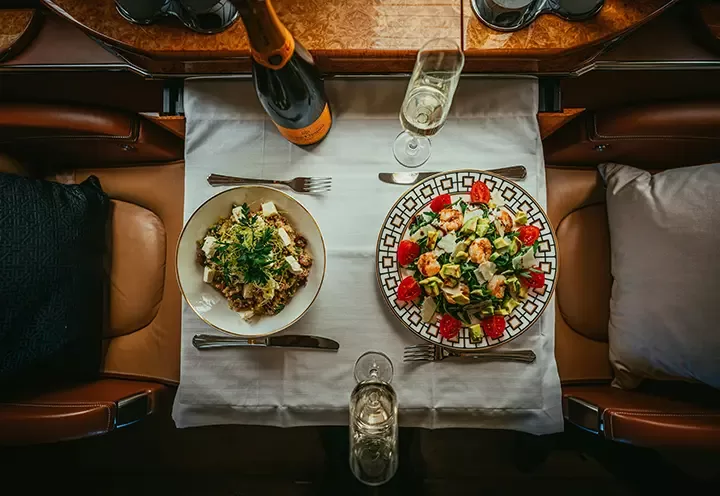 Aerial view of a table set for two with a bottle of champagne, two filled glasses, a bowl of pasta with herbs, and a plate of salad with tomatoes and greens on a white tablecloth.