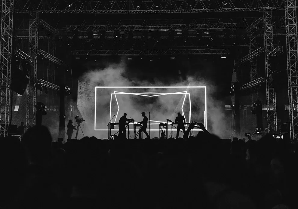A black and white photo of a live concert with silhouetted musicians performing on stage. Bright geometric lines illuminate the backdrop, and the audience is partially visible in the foreground.