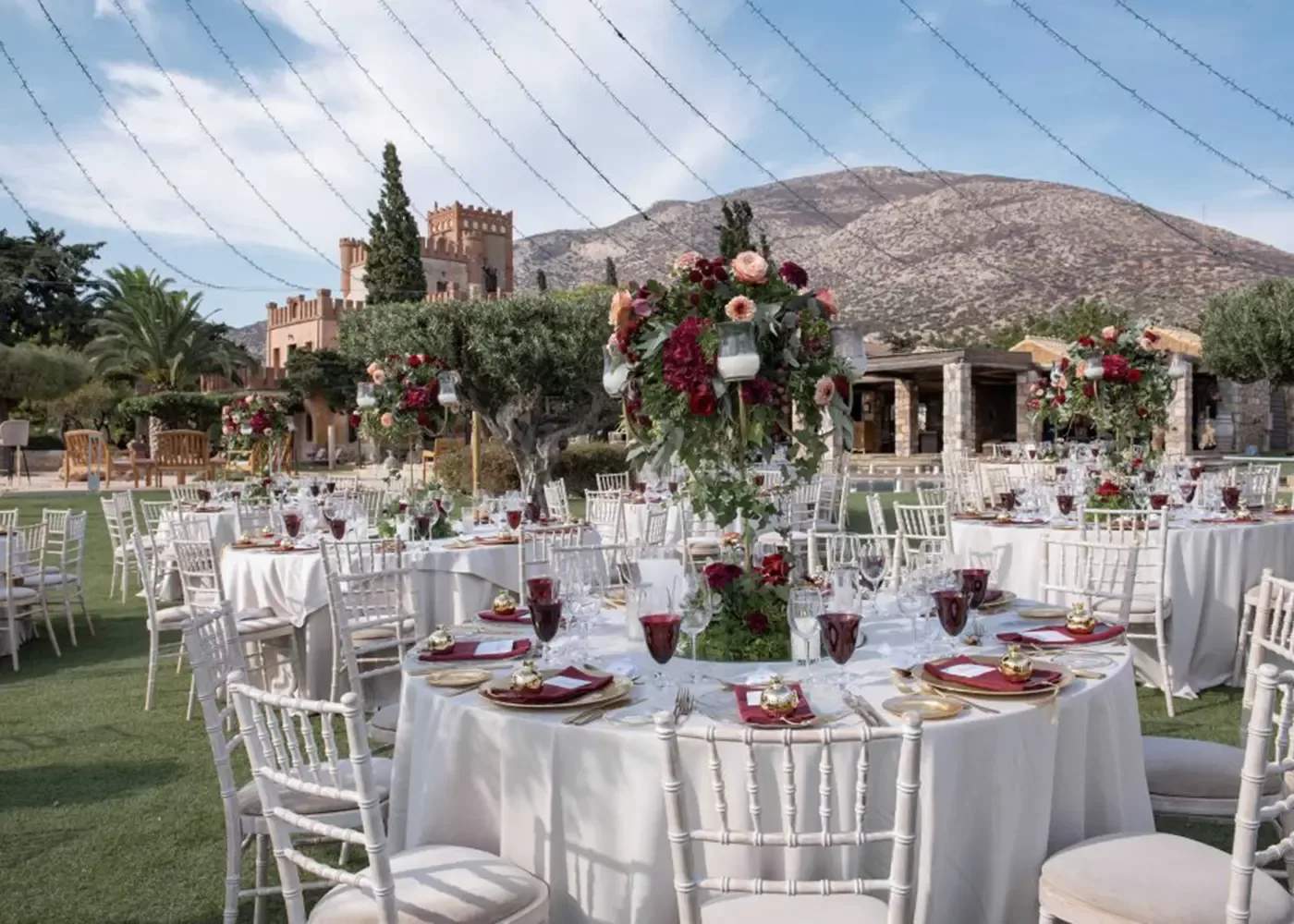 An outdoor wedding reception with round tables covered in white tablecloths and decorated with tall floral centerpieces. Elegant place settings feature red goblets and gold accents. In the background, a castle-like building and mountains are visible.