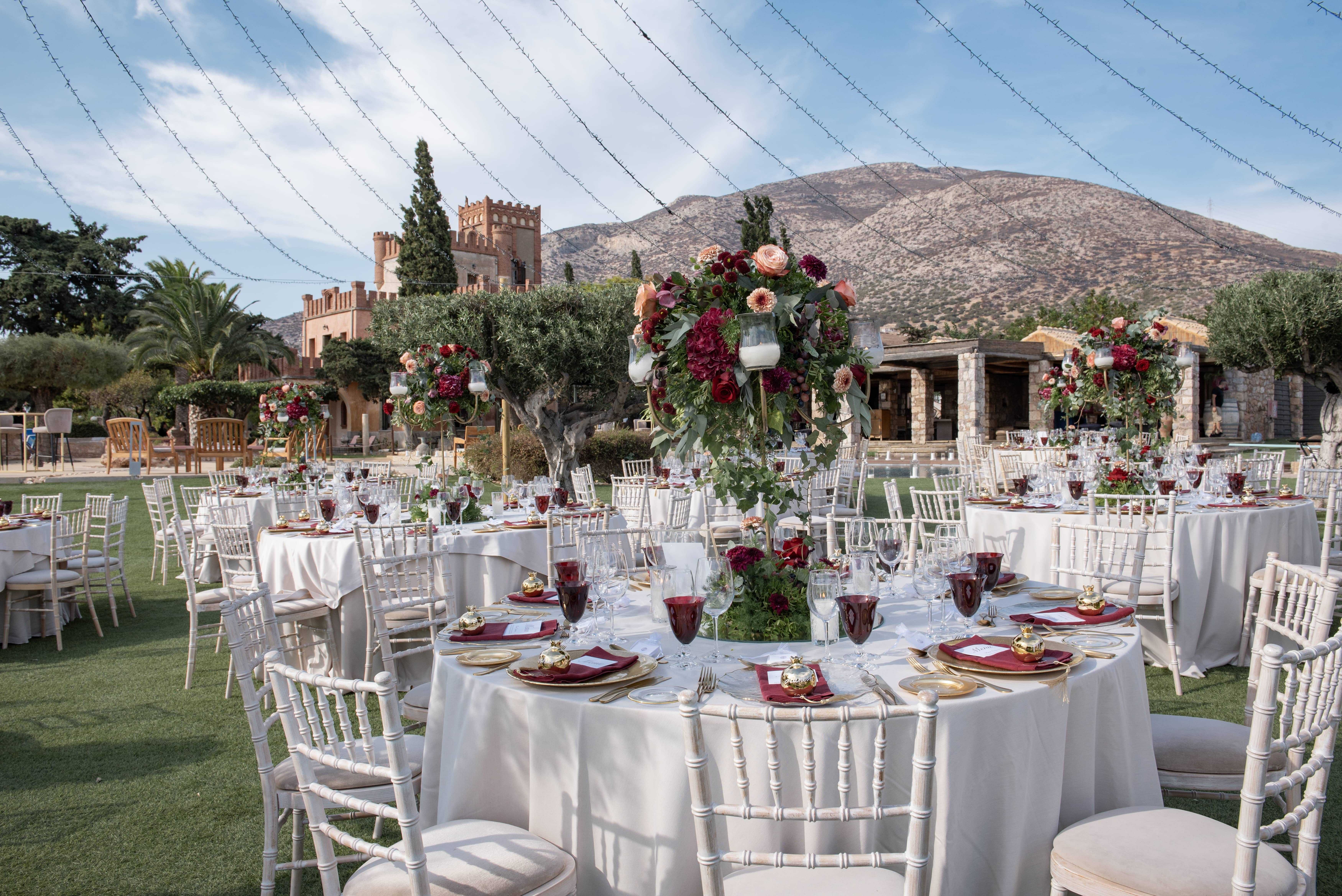 An outdoor wedding reception with round tables covered in white tablecloths and decorated with tall floral centerpieces. Elegant place settings feature red goblets and gold accents. In the background, a castle-like building and mountains are visible.