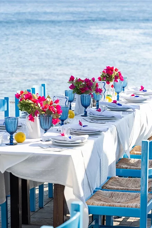 A long outdoor dining table by the sea, covered with a white tablecloth. Blue chairs surround the table, which is decorated with blue glasses, white plates, pink flower arrangements, and lemons.