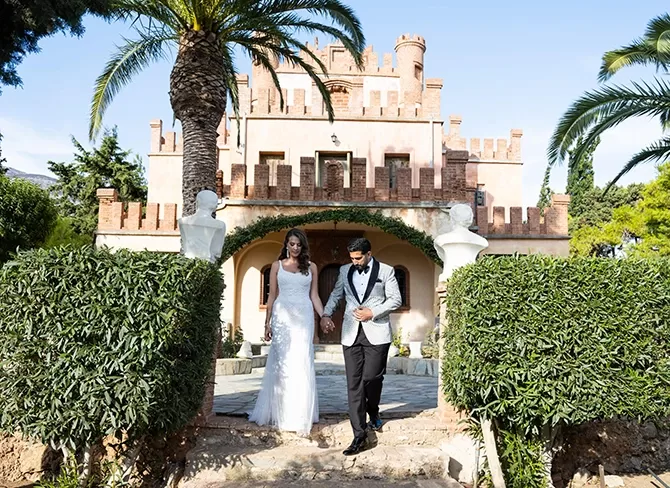 A couple in wedding attire walks hand in hand down a stone path, surrounded by lush greenery. Behind them stands a grand, castle-like building with a palm tree nearby, under a clear blue sky.