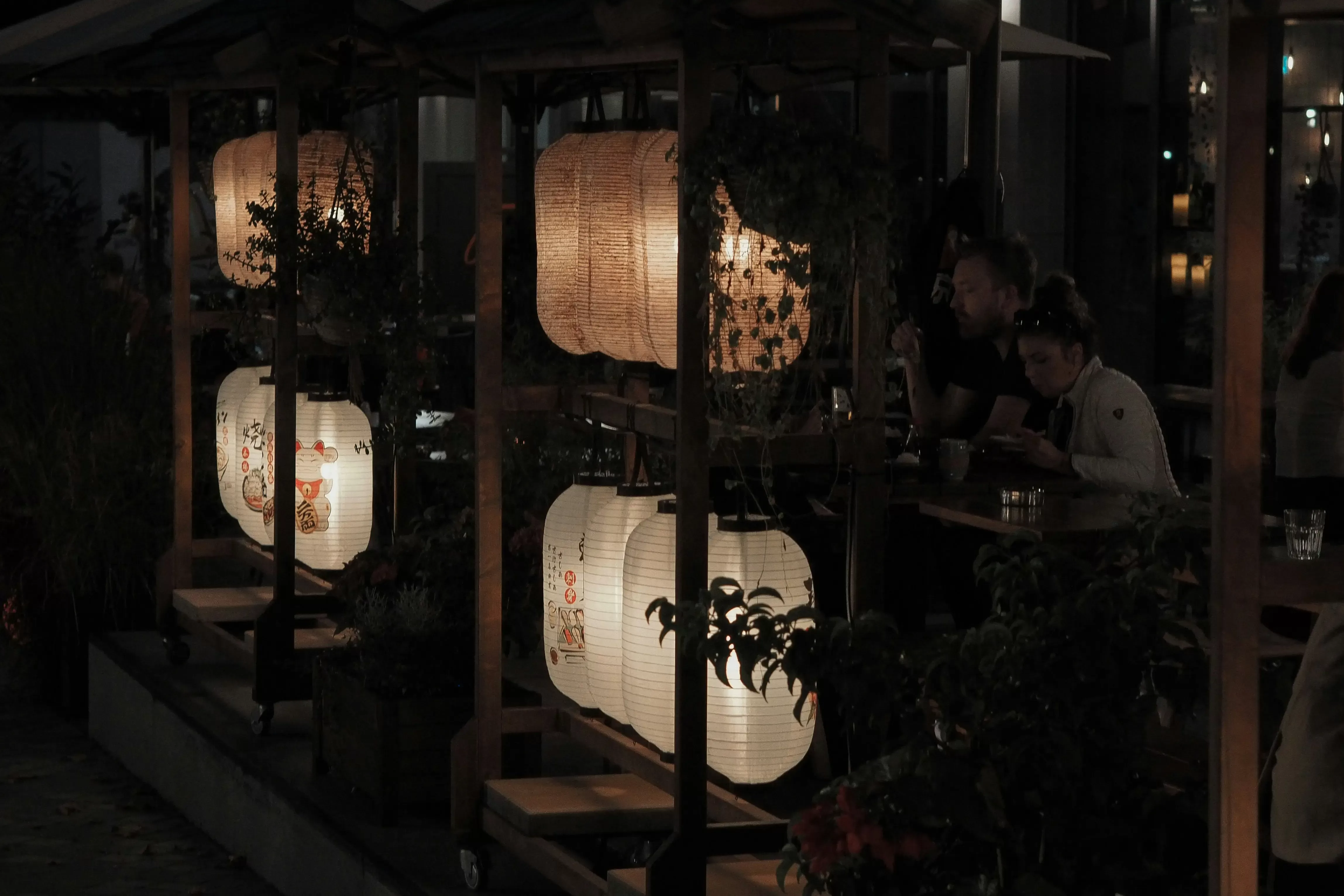 People dine at outdoor tables in a dimly lit setting, decorated with glowing Japanese paper lanterns hanging from wooden structures, creating a cozy and intimate nighttime atmosphere.