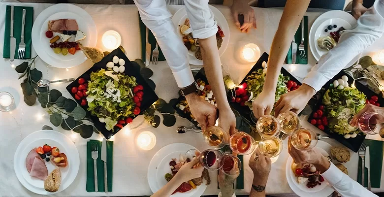 Areal view of a  group of individuals hands joyfully toasting with glasses at a beautifully set dinner table filled with food.