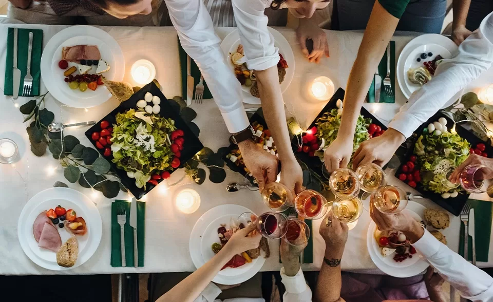 Areal view of a  group of individuals hands joyfully toasting with glasses at a beautifully set dinner table filled with food.