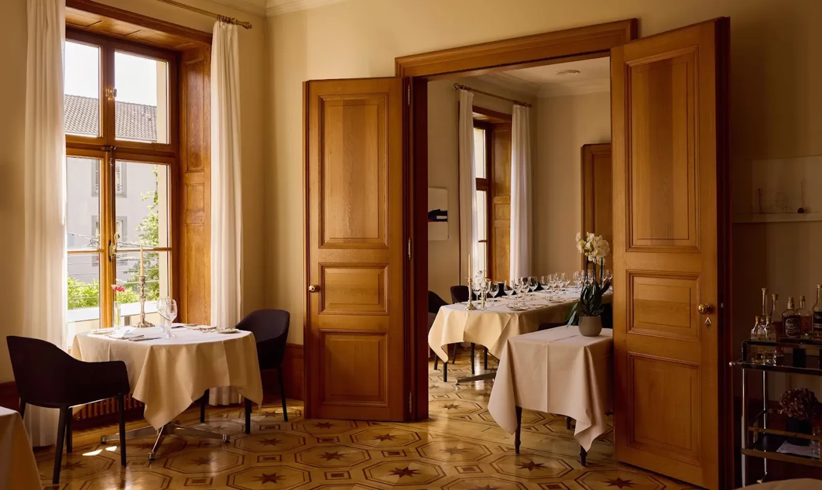 A cozy, elegant restaurant dining area with wooden doors, large windows, round tables covered with white tablecloths, dark chairs, and sunlight streaming in across patterned wood floors.