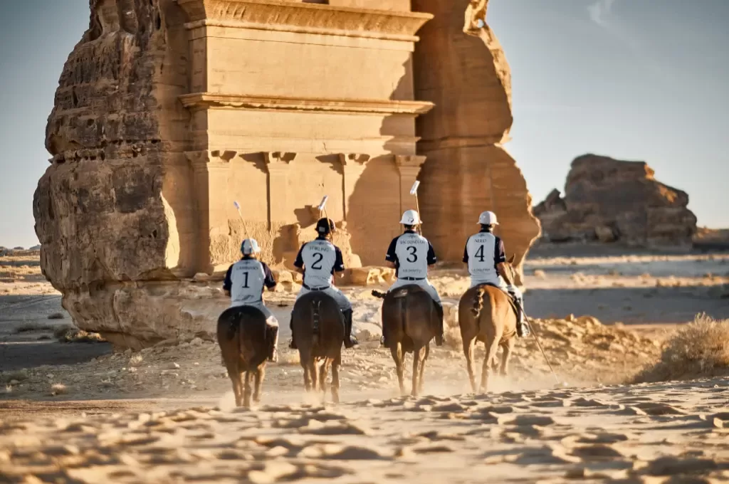 Back view of four male polo players riding on horses in uniform numbered shirts in the desert