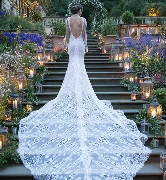 Bride in beautiful lace backless wedding dress stood on stairs with lights and floral decor with her train draping down the staircase. | Quintessentially Weddings