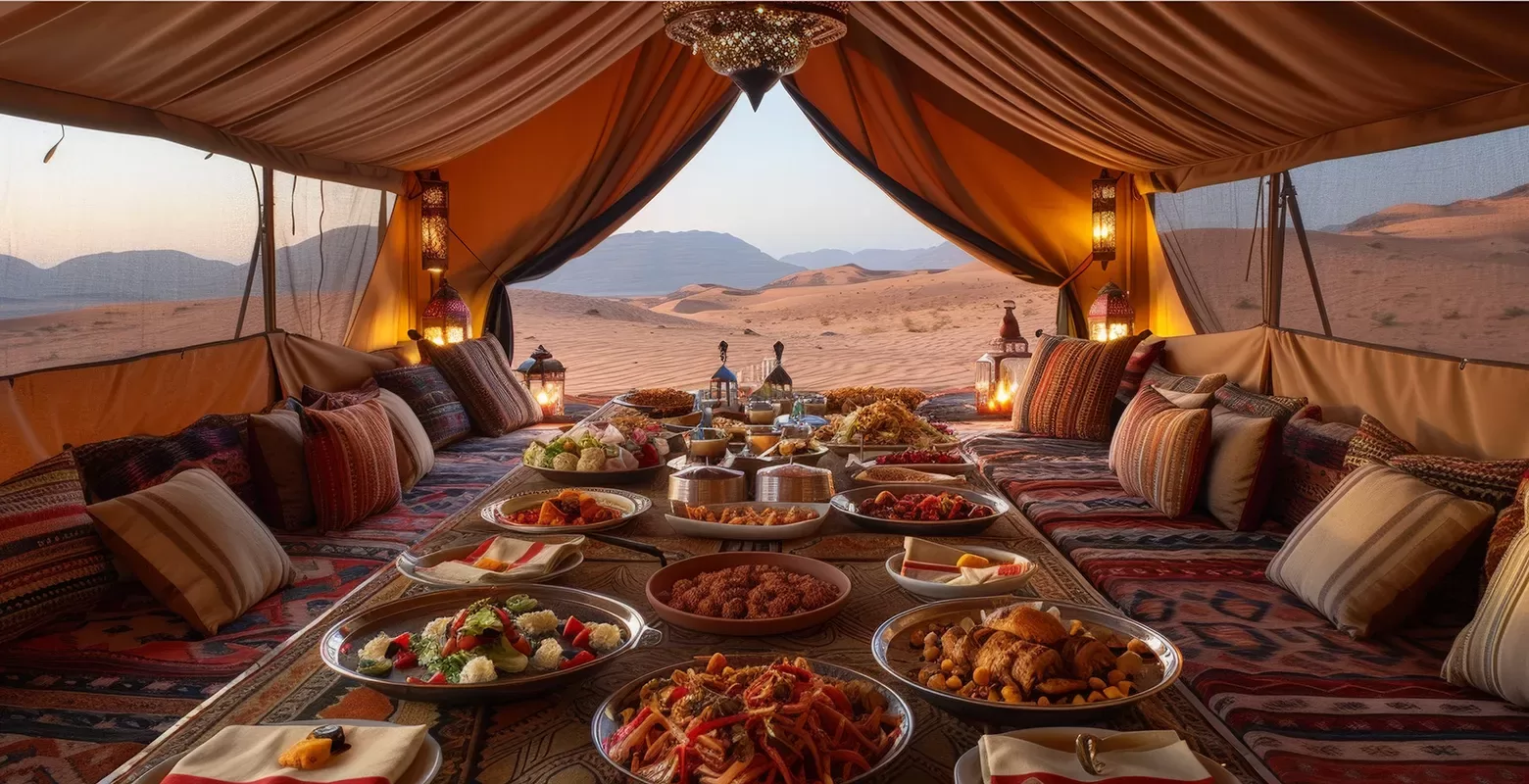 A lavish spread of Middle Eastern dishes is laid out on a low table within a tent in the desert. The tent is adorned with patterned cushions and lanterns, offering a view of sand dunes and a mountainous horizon at sunset.