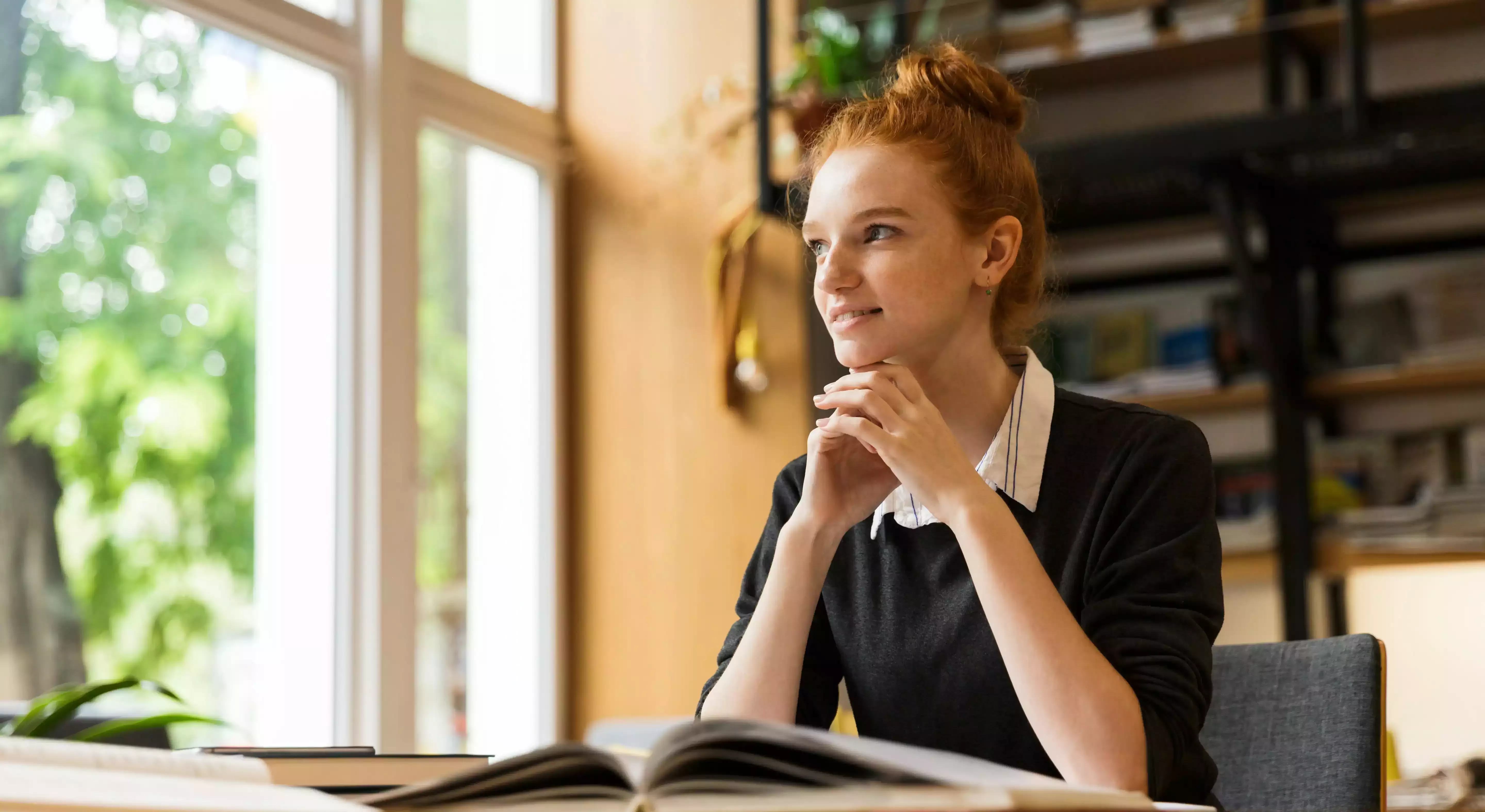 Female student sat at desk with a book smiling looking outside the window