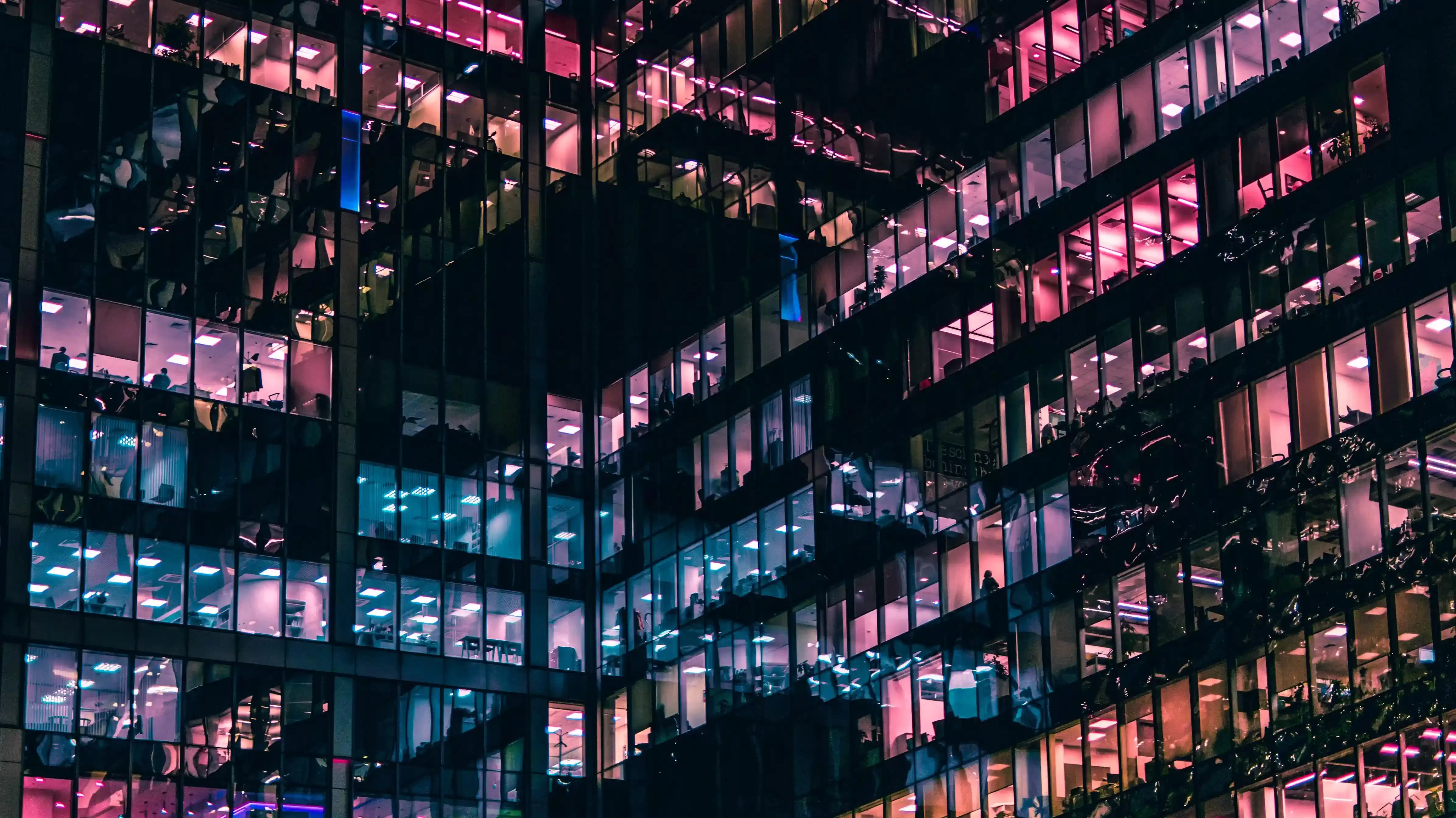 Photo of exterior of fully glassed office building with various pink, blue, orange and purple lights