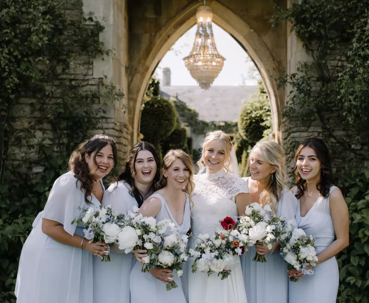 A bride and five bridesmaids smile while holding floral bouquets. They stand under a stone arch adorned with greenery, with a chandelier overhead. The bridesmaids wear light blue dresses, and the setting is an outdoor garden area.