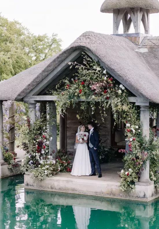 A newlywed couple stands under a rustic, thatched-roof gazebo adorned with lush greenery and colorful flowers, overlooking a pool of turquoise water. The bride holds a bouquet, and both gaze at each other lovingly.