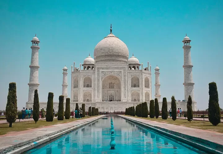 The Taj Mahal, a white marble mausoleum with a large dome, four minarets, and intricate carvings, stands reflected in a long rectangular pool under a clear blue sky. Visitors walk along the pathway lined with trees.