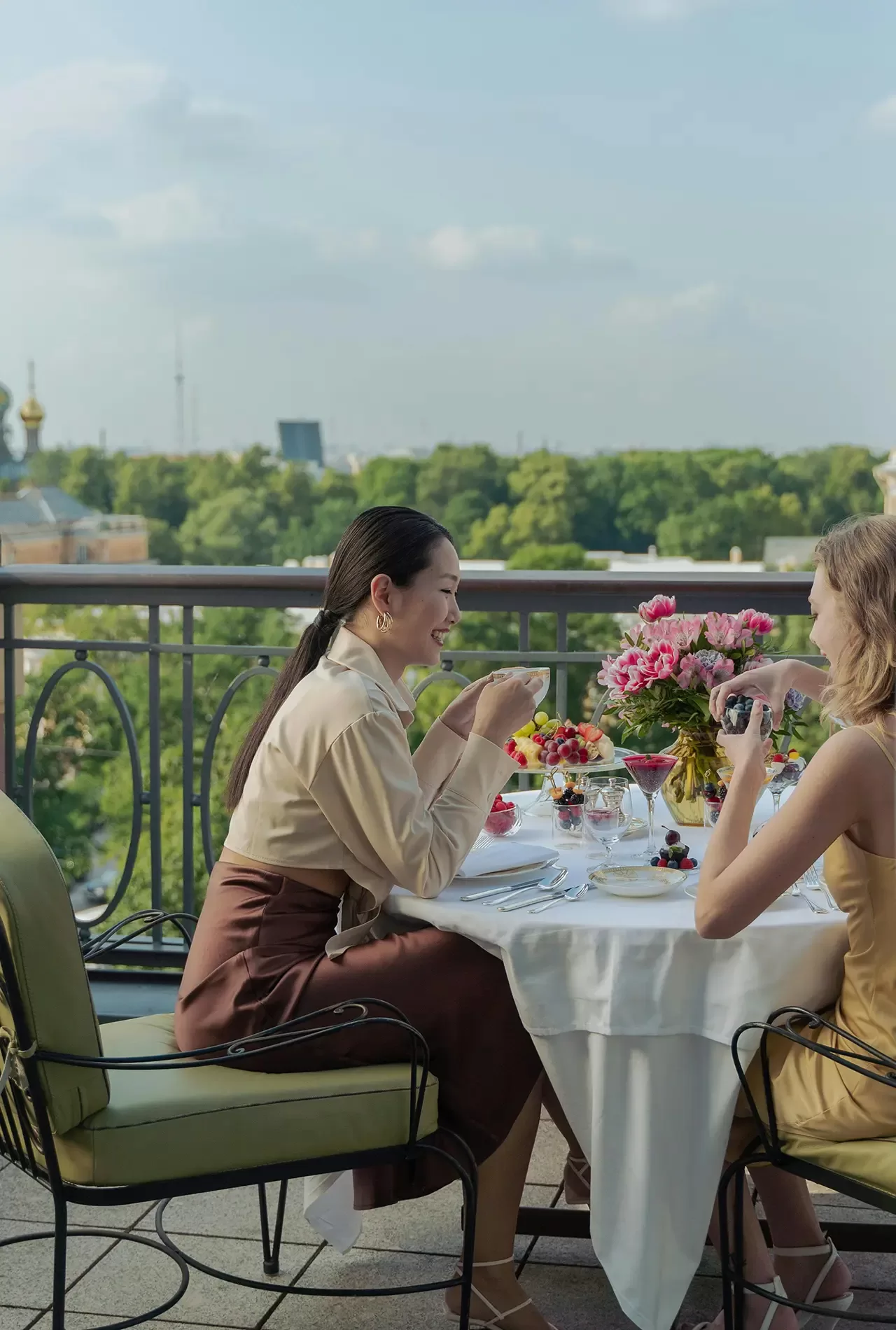 Two women sit at an outdoor table, enjoying food and drinks on a balcony with a scenic view of trees and distant buildings. The table is decorated with flowers and elegant place settings.