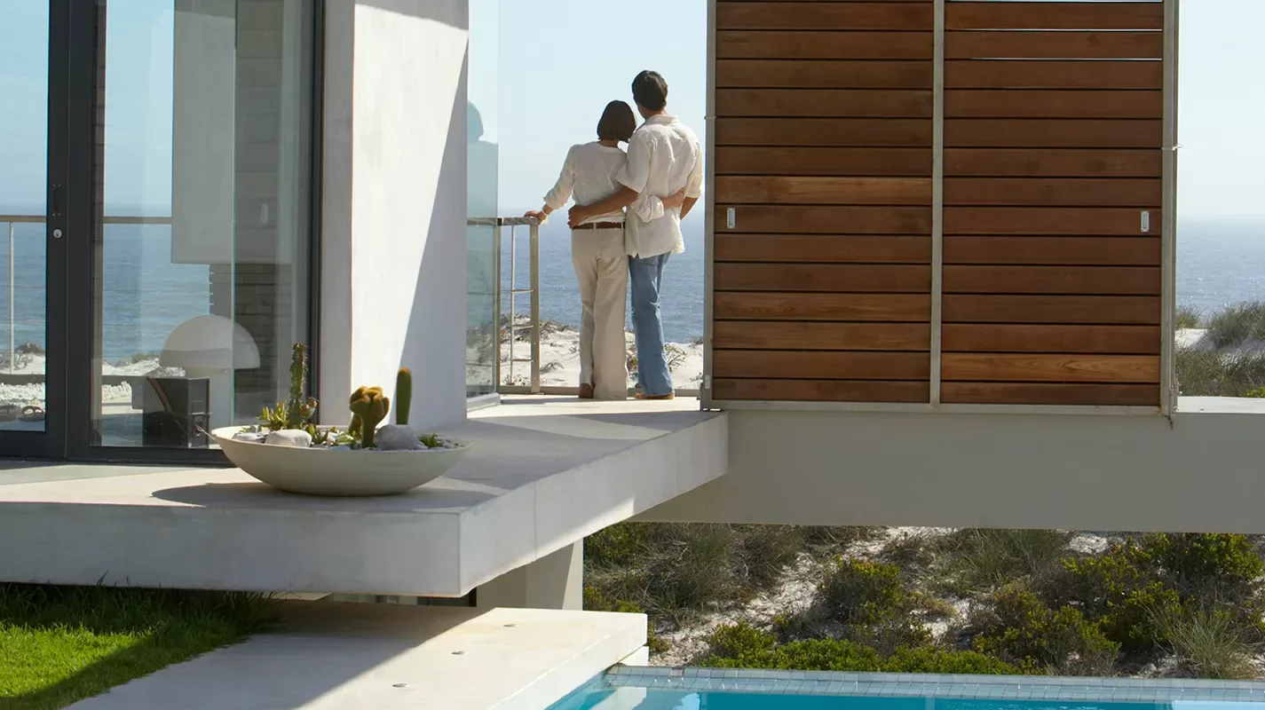 A couple stands arm in arm on a modern balcony, looking out at the ocean. The home features clean lines, a pool below, and a desert landscape with sand and sparse vegetation in the background.