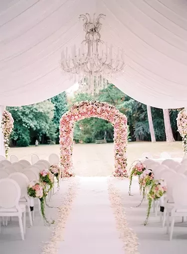 Beautiful white interior wedding ceremony set up with pink flower decors, glass chandelier under white draped roof | Quintessentially Weddings