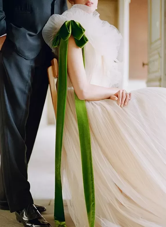 Women in a beautiful avant-garde wedding dress with velvet green bow shoulder detail sat in front of a groom in a suit.