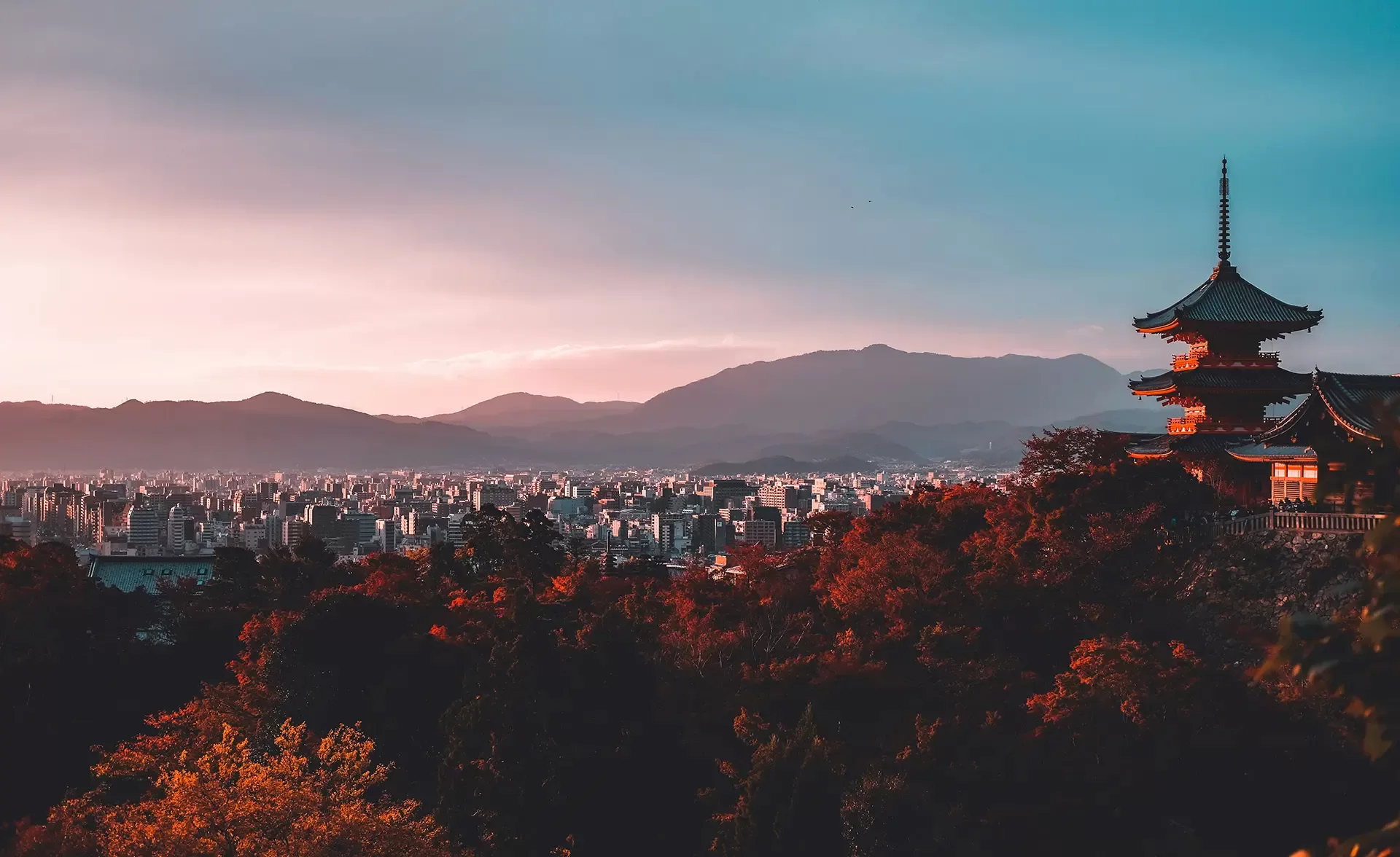 A serene landscape featuring a traditional pagoda on the right, surrounded by autumn-colored trees. In the background, a sprawling city under a soft, pastel sky with mountains silhouetted in the distance.