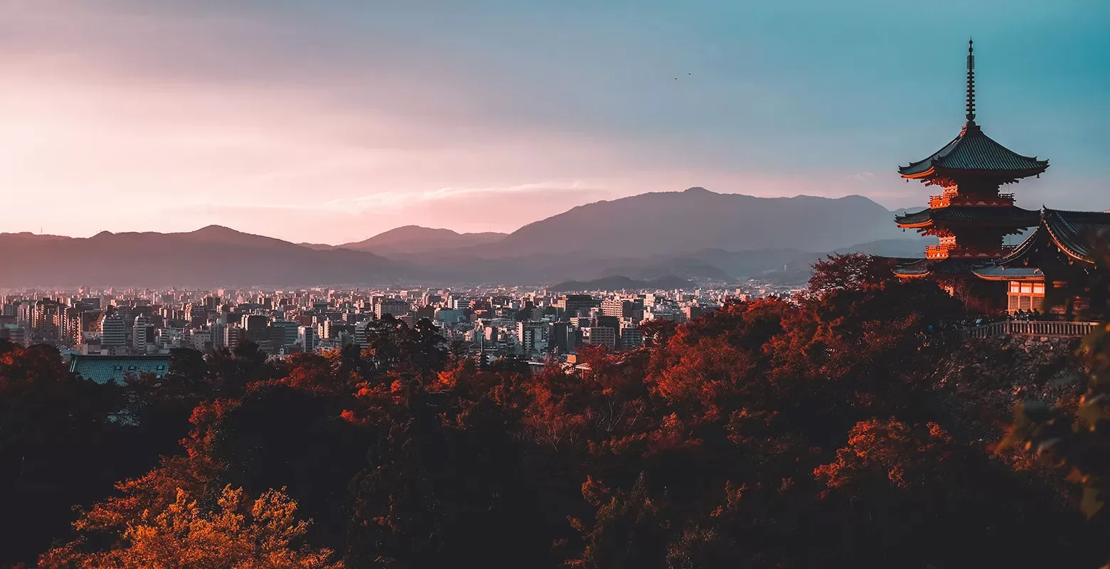 A serene landscape featuring a traditional pagoda on the right, surrounded by autumn-colored trees. In the background, a sprawling city under a soft, pastel sky with mountains silhouetted in the distance.