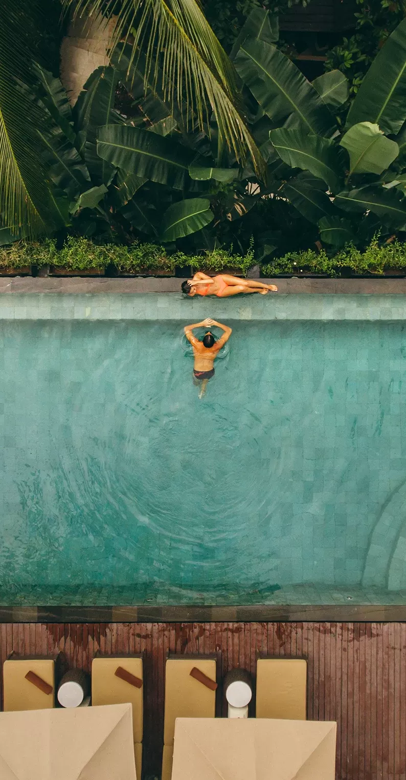 Birdseye view of outdoor swimming pool and loungers, amongst huge green forest trees with a women bathing on the edge of the pool and a man inside of the pool.