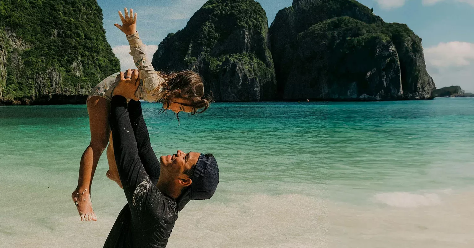 A man wearing a dark shirt and cap lifts a smiling young girl above his head on a sunny beach, with turquoise water and lush green cliffs in the background.