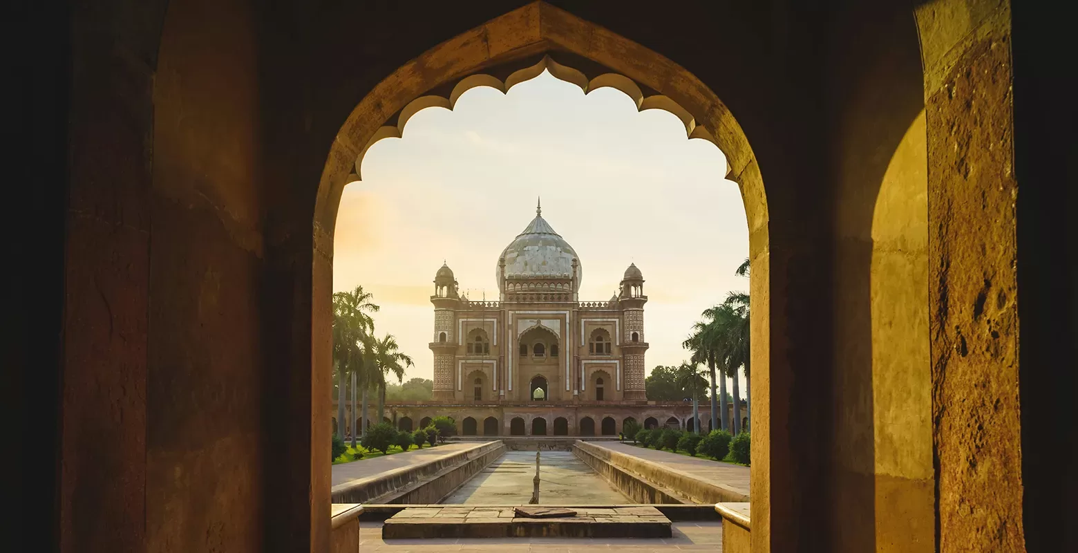 A view of Humayun’s Tomb in Delhi, India, framed by an ornate arched doorway, with the mausoleum’s domed structure and surrounding palm trees visible at sunset.