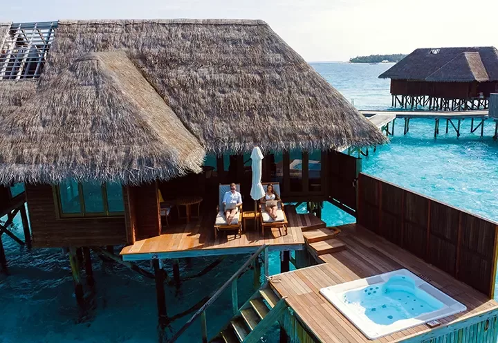 A couple relaxes on lounge chairs on the deck of an overwater bungalow, with a thatched roof, above clear blue ocean. A jacuzzi is on the deck, and another bungalow is visible in the background.
