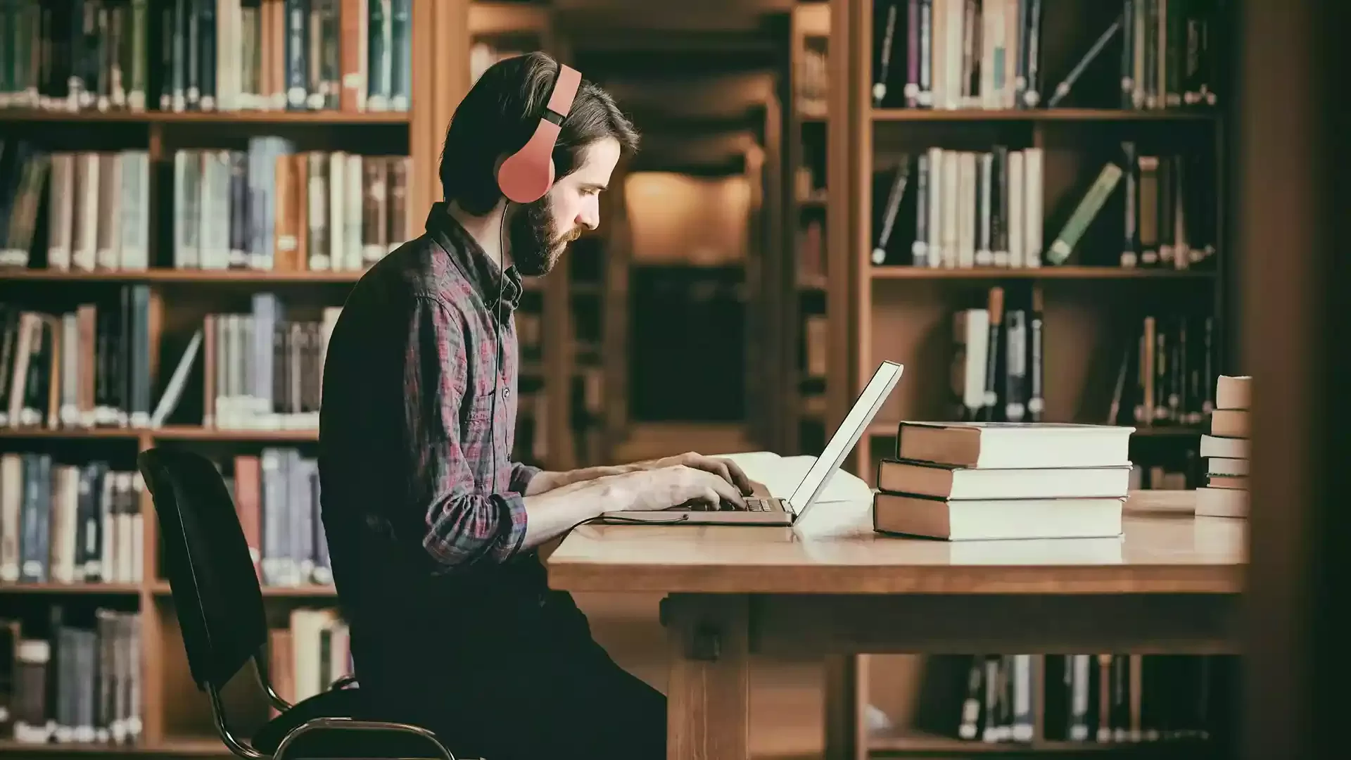 Man wearing headphones sat at a desk with a stack of books and a laptop in a library