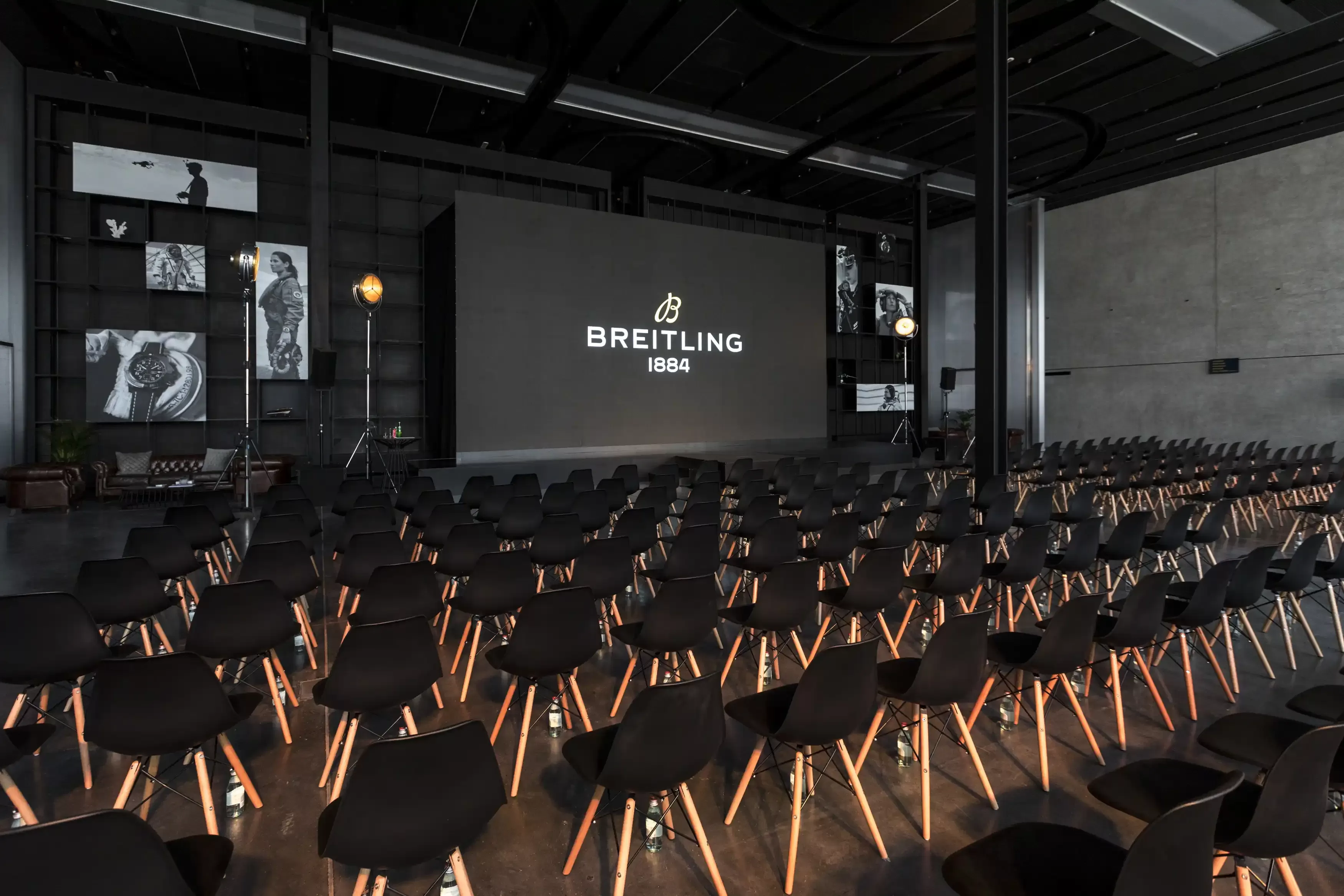 Black themed set up of rows of black chairs with wooden legs facing a stage with Breitling projector adjacent to black and white gallery of images