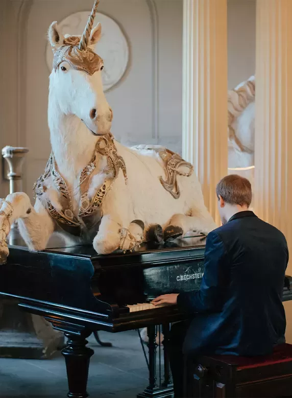 A male pianist sat playing a grand piano which has a large white and gold unicorn sculpture sat on top of the pano.