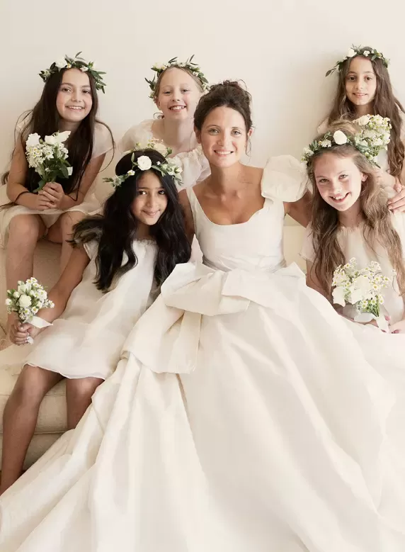 Bride in a wedding dress with bow detail sat with young bridesmaids holding flowers and wearing flower headpieces.