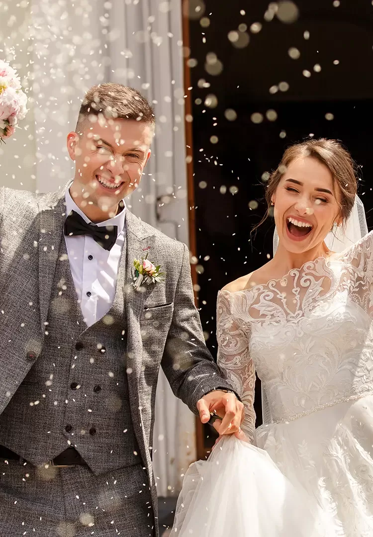 A joyful couple celebrates their wedding. The groom, in a gray suit and bow tie, and the bride, in an elegant white lace dress, hold hands and smile as confetti falls around them outside a building.