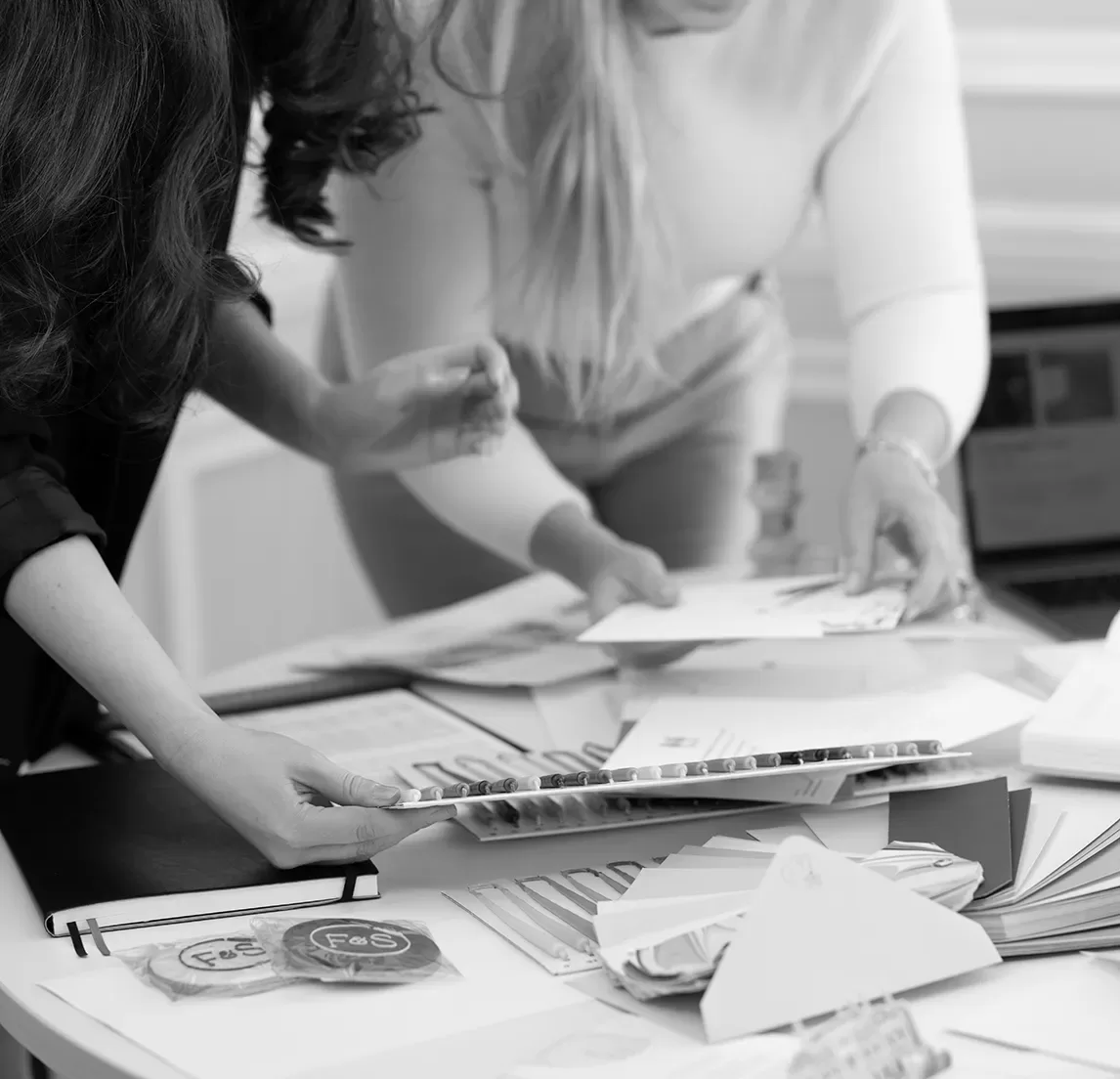 Two people are gathered around a table covered with papers, envelopes, and color samples. They appear to be discussing and organizing materials. A laptop is partially visible in the background. The scene is in black and white.