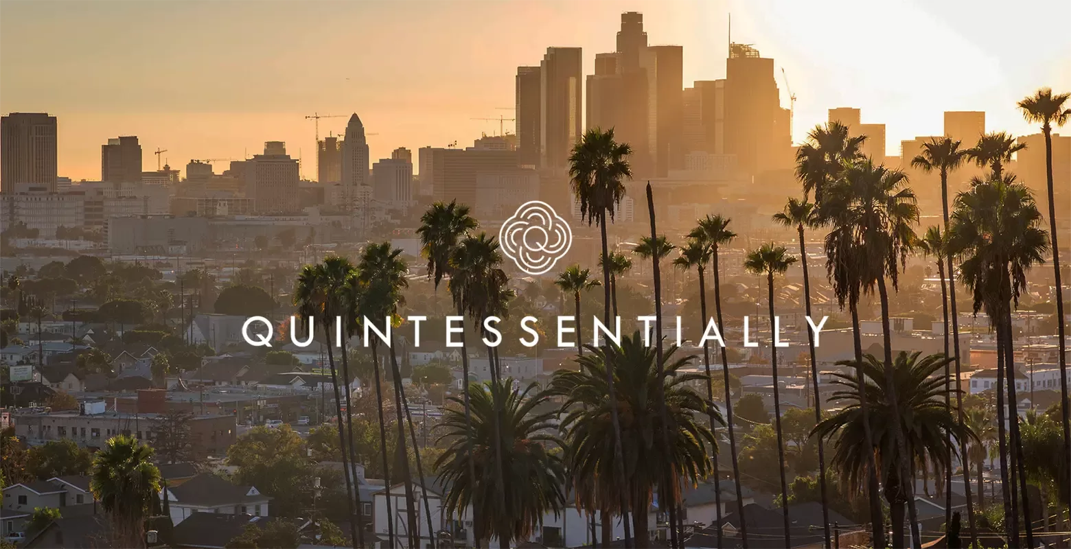 A cityscape of Los Angeles at sunset with palm trees in the foreground, skyscrapers in the background, and the word QUINTESSENTIALLY with a floral logo in the center.