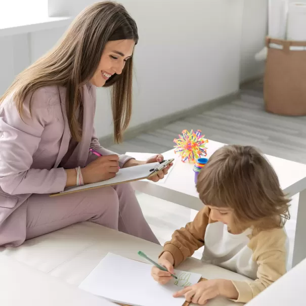 Women in pink suit with notepad smiling down at child writing on paper on a bench