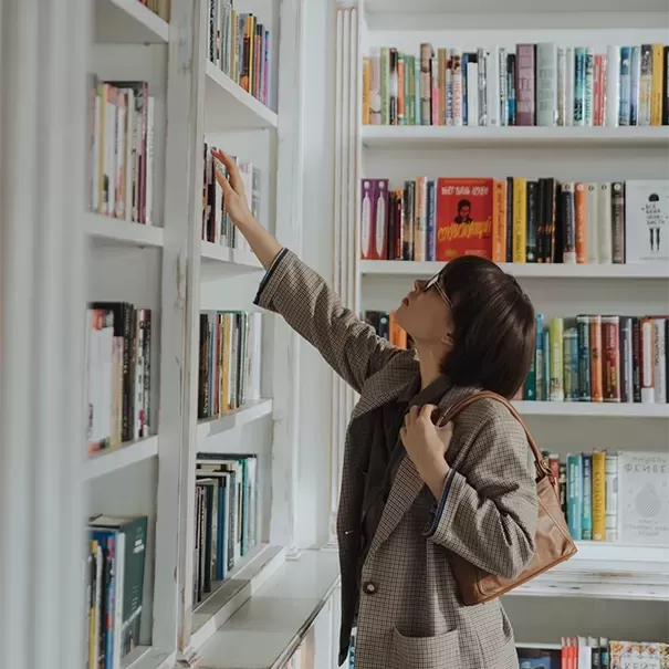 Women in a library reaching up to a book on a white book shelf