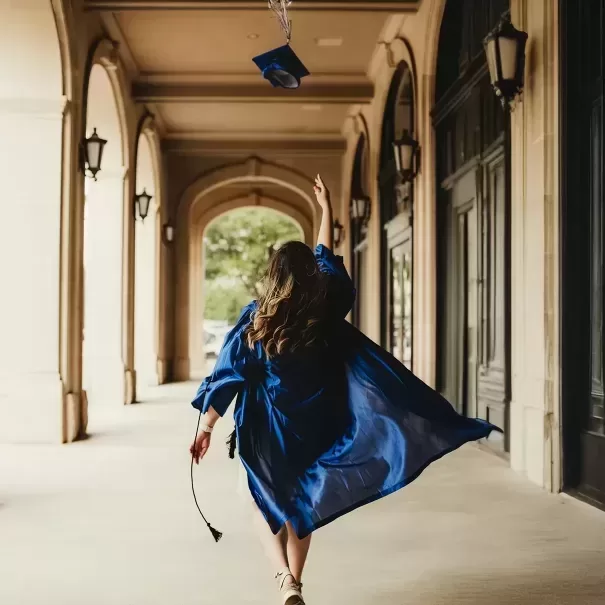 A person in a blue graduation gown tosses their cap into the air while walking down an arched corridor, holding a diploma in one hand.