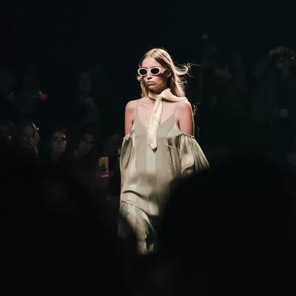 A model walks confidently on the runway wearing a pale green off-shoulder dress, a light-colored scarf, and white sunglasses. The background is dimly lit with an audience watching and capturing the event.