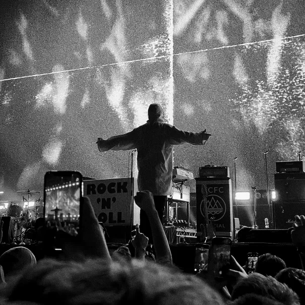 A performer stands on stage with arms outstretched facing a cheering crowd. Audience members hold up phones to record. The stage is lit with dramatic lights and equipment, and a Rock N Roll sign is visible. The image is in black and white.