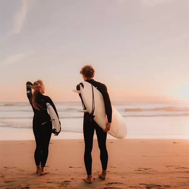 Man and women in wetsuits, carrying surfboards standing in the sand overlooking the sea at sunset