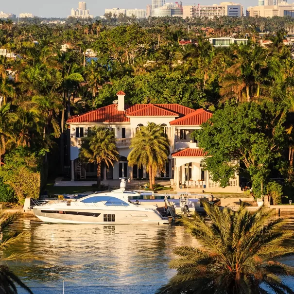 Elegant waterfront mansion with a red-tiled roof, surrounded by lush palm trees. A luxury yacht is moored in front, and the background features a city skyline bathed in the golden glow of sunset.